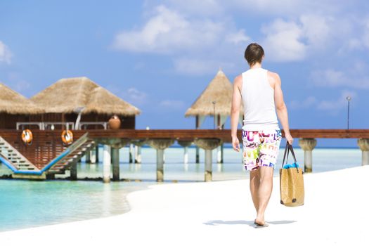 Man walking by the shore in the beach going away to a jetty with bungalows. He is wearing a colorful bathing suit while carrying a beach bag with towels. At background there are bungalows out of focus.