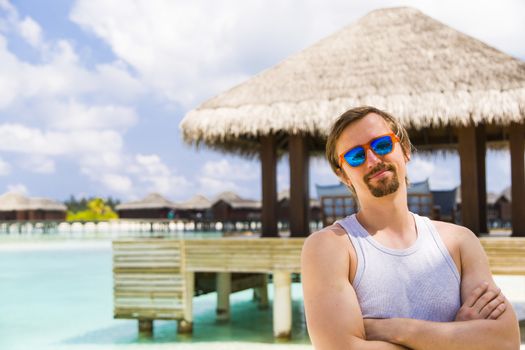 Young man looking at the camera with his arms crossed. He is wearing a blue mirror sunglasses, he is smiling. At background there are bungalows over turquoise clear water.