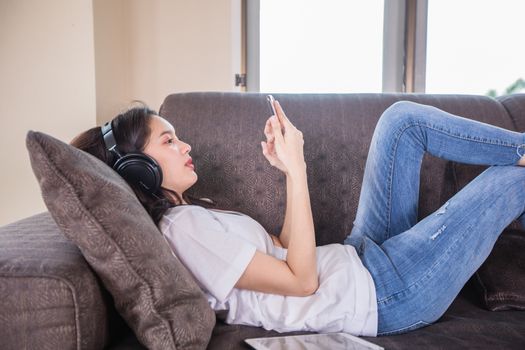 Asian young woman happy cheerful cute beautiful wears a headset for listening to music from a mobile phone and she is playing smartphone and lying on the sofa at home,