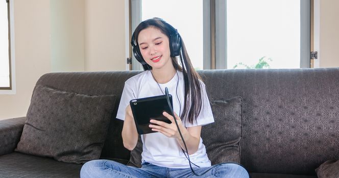 Asian young woman happy cheerful cute beautiful wears a headset for listening to music from a tablet and she is playing tablet and sitting on the sofa at home,