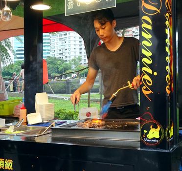 KAOHSIUNG, TAIWAN -- JUNE 9, 2016: A young male chef sears pieces of steak with a blowtorch at an outdoor food stall.
