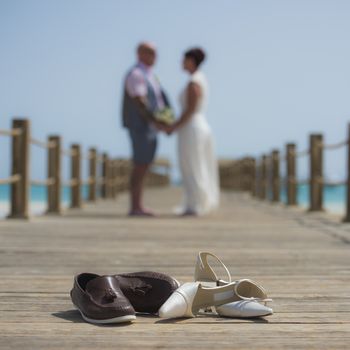 Married couple on wedding day walking romantically outside on wooden pier with pair of shoes in foreground