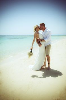 Beautiful married couple kissing at a tropical beach paradise on wedding day in white gown dress with ocean view vintage style photo