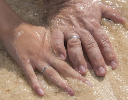 Hands of bride and groom on wedding day showing rings at tropical sandy beach in water romantic concept