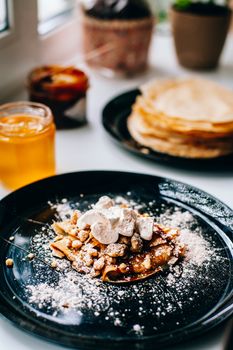 Pancakes with caramel nuts and marshmallows on the kitchen windowsill. In the background are a lot of pancakes, caramel and honey in a jar.