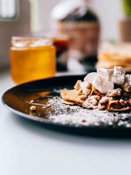 Pancakes with caramel nuts and marshmallows on the kitchen windowsill. In the background are a lot of pancakes, caramel and honey in a jar.