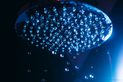 Drops of water fall from a watering can in the shower in blue light. Water drops close-up. Flow of water. Big round watering can macro photo.