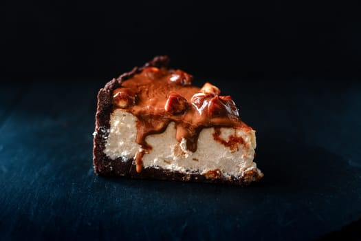 A piece of cheesecake with hazelnuts and caramel on a black background.