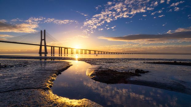 Vasco da Gama bridge at sunrise in Lisbon, Portugal. Vasco da Gama Bridge is a cable-stayed bridge flanked by viaducts and rangeviews that spans the Tagus River in Parque das Nações in Lisbon. 