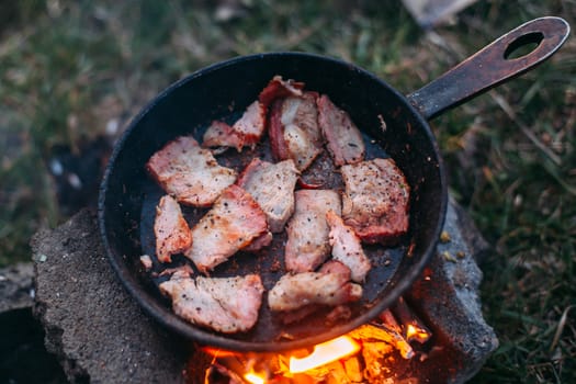 Slices of fried bacon in a pan. Food in a forest camp. Cooking on fire. Picnic in the nature. Grilled food on nature.