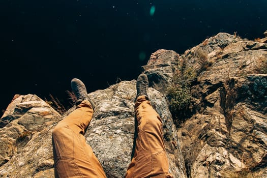 Man sitting on a rock. Photo of feet on a background of water. A guy in brown pants on the edge of a cliff.
