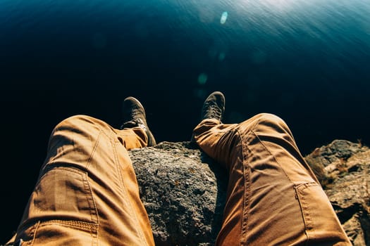 Man sitting on a rock. Photo of feet on a background of water. A guy in brown pants on the edge of a cliff.