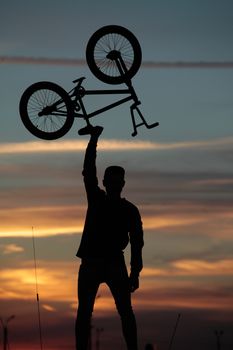 Silhouette of a cyclist holding his bike over his head on a sunset background, standing by a car