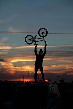 Silhouette of a cyclist holding his bike over his head on a sunset background, standing by a car
