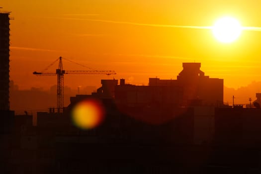 Orange sunset on the background of the city. Silhouette of a construction crane. Photo with a glare from the sun. Panorama of the city.