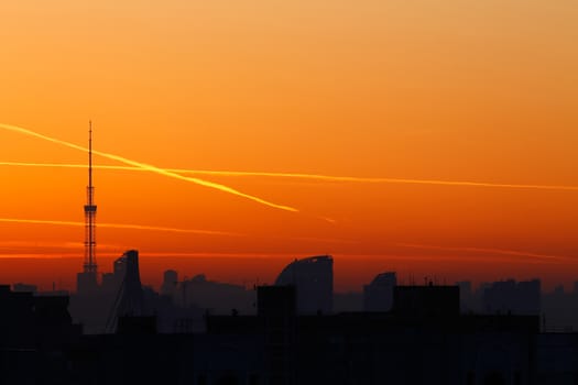A television tower but against a background of bright orange sunset and the city. Traces of the plane in the sunset sky.