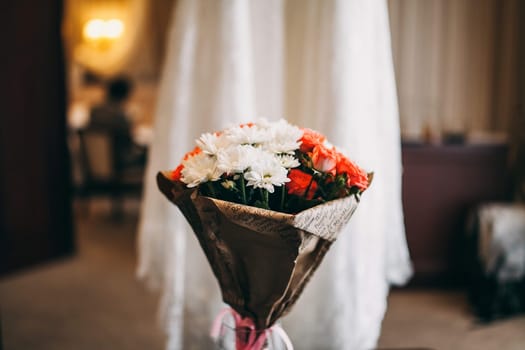 Bouquet of pink roses and white asters standing in a vase on a black glass table on the background of which a wedding dress