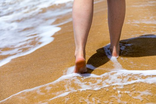 Woman walking barefoot on a beach. Close up leg of young woman walking along wave of sea water and sand on the summer beach. Travel Concept. Woman walking on sand beach leaving footprints in the sand.