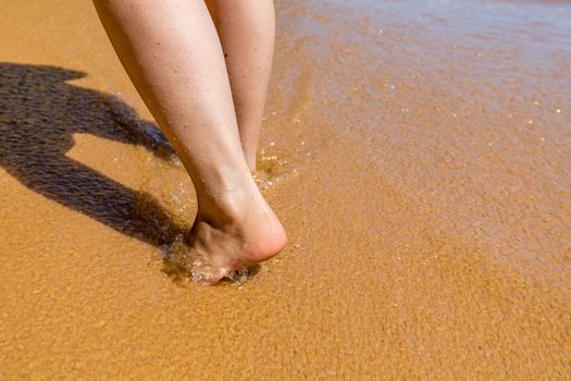 Woman walking barefoot on a beach. Close up leg of young woman walking along wave of sea water and sand on the summer beach. Travel Concept. Woman walking on sand beach leaving footprints in the sand.