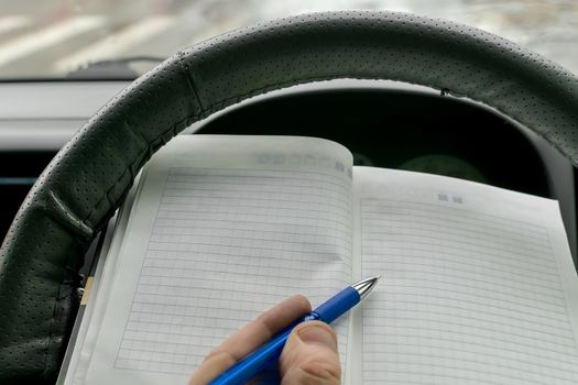 the hand of the car driver behind the wheel holds a paper notebook and makes notes