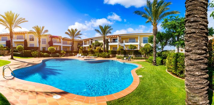 Swimming pool, sun-loungers and palm trees during a warm sunny day, paradise destination for vacations. Backyard swimming pool with garden full of palm trees and flowers. Backyard with swimming pool.