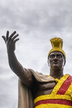 Kapaau, Hawaii, USA. - January 15, 2020: Closeup of head and stretched arm of statue of King Kamehameha, decorated with yellow and red dress, golden crown under gray cloudscape.