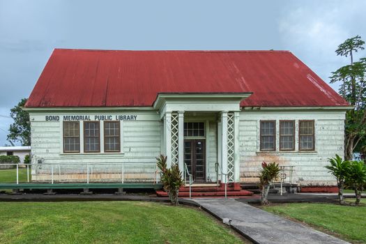 Kapaau, Hawaii, USA. - January 15, 2020: Historic wooden, light green walls, and red roof, Bond memorial public library set on green lawn under blueish sky, Green foliage on sides,