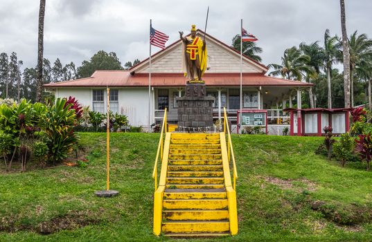Kapaau, Hawaii, USA. - January 15, 2020: Yellow stairway to and statue of King Kamehameha, decorated with yellow and red dress, golden crown and long spear under gray cloudscape with US and Hawaii flags.