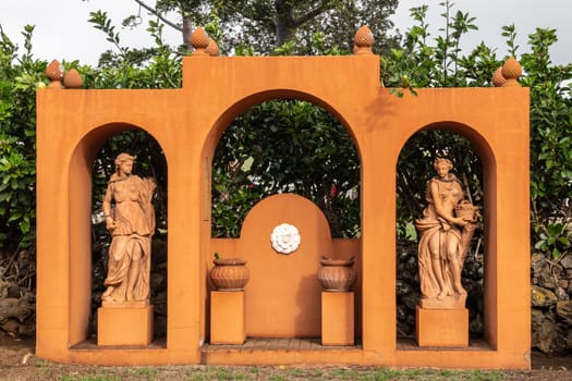 Waimea, Hawaii, USA. - January 15, 2020: Parker Ranch headquarters. Orange statue in garden showing three bows whereunder two women and pots. Green foliage.
