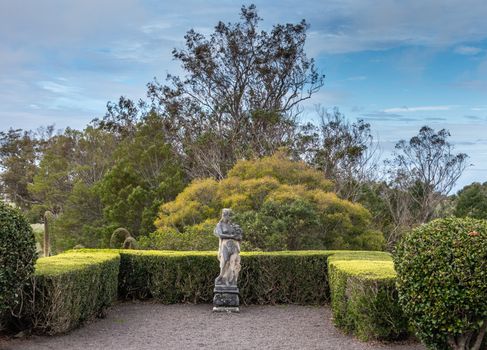 Waimea, Hawaii, USA. - January 15, 2020: Parker Ranch headquarters. Old white stone discollored by mold statue of half-nude woman with grapes under blue sky. Green foliage in back.