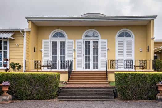 Waimea, Hawaii, USA. - January 15, 2020: Parker Ranch headquarters. Yellow walls with white window frames and shutters of wing of main house. Green hedge and stairs in fornt. Silver sky.