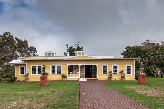 Waimea, Hawaii, USA. - January 15, 2020: Parker Ranch headquarters. Main house is painted yellow with white framed door and windows under heavy cloudscape. Green foliage and old statues.