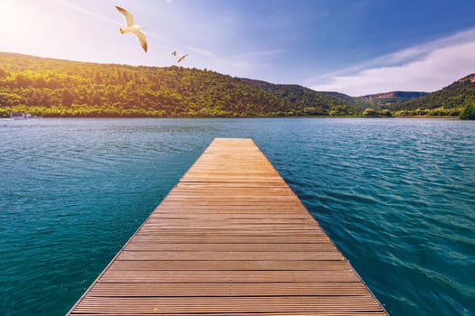 Idyllic view of the wooden pier in the lake with mountain scenery background. Wooden bridge on the lake. Long pier on lake and blue sky in summer.  Seagulls over pier.