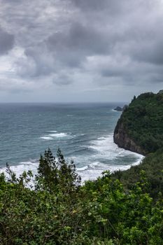 Kohala, Hawaii, USA. - January 15, 2020: Portrait of Coastline with white surf where Pololu valley and its beach meets the ocean. Tall steep cliffs, green forests under heavy dark rain cloudscape.