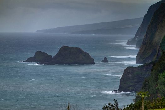 Kohala, Hawaii, USA. - January 15, 2020: Coastline where Pololu valley and its water meets the ocean. Boulder islands, tall steep cliffs, all under heavy rain cloudscape. Some green foliage.