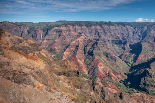 Waimea Canyon, Kauai, Hawaii, USA. - January 16, 2020: Wide landscape with waterfall in distance shows red rocks partly green in crevasses, green cover on top, under blue sky.