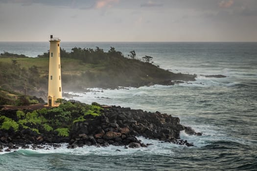 Nawiliwili, Kauai, Hawaii, USA. - January 16, 2020: Yellow Ninini lighthouse on black ocean-shore rocks early morning. Dark gray ocean under light gray sky. Some green foliage on land.