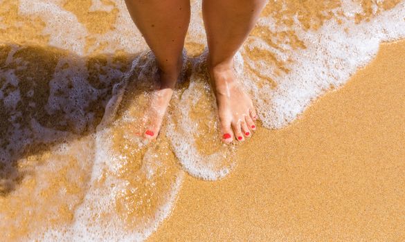 Closeup on leg of young woman standing on sea shore. Woman legs and feet walking on the sand of the beach with the sea water in the background. Woman standing on the beach with wave sea.
