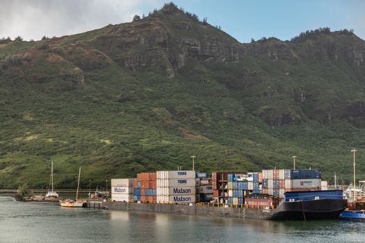 Nawiliwili, Kauai, Hawaii, USA. - January 17, 2020: Full Haaheo shipping container barge docked in port. Green mountain with brown-black cliffs as backdrop under light blue cloudscape.