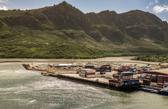 Nawiliwili, Kauai, Hawaii, USA. - January 17, 2020: Almost empty Haaheo shipping container barge docked in port. Green mountain with brown-black cliffs as backdrop under light blue cloudscape.