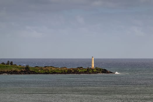 Nawiliwili, Kauai, Hawaii, USA. - January 17, 2020: Yellow Ninini lighthouse on black ocean-shore rocks of its green lands end. gray ocean water and light blue cloudscape.