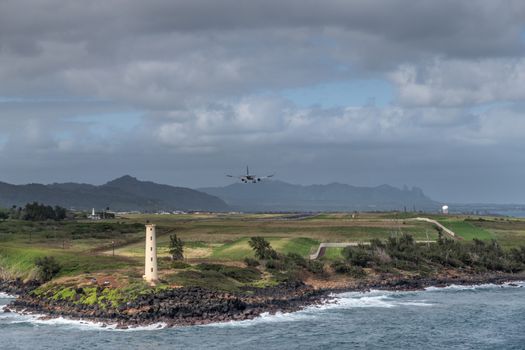 Nawiliwili, Kauai, Hawaii, USA. - January 17, 2020: Airplane lands at airport behind Yellow Ninini lighthouse on black ocean-shore rocks. Blueish ocean water and heavy dark cloudscape.