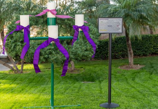 Garden Grove, California, USA - December 13, 2018: Crystal Christ Cathedral. Closeup of the Advent Wreath in the garden with explaining poster. Green backgroound, four white candles, purple and pink ribbons.