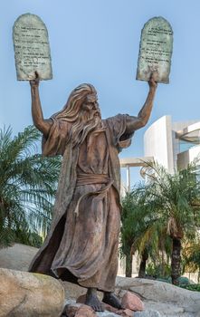 Garden Grove, California, USA - December 13, 2018: Crystal Christ Cathedral. Bronze statue of Moses putting the ten commandments of two tables in the air. Some green foliage, blue sky, white Cultural Center.