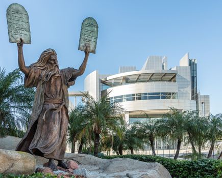 Garden Grove, California, USA - December 13, 2018: Crystal Christ Cathedral. Bronze statue of Moses putting the ten commandments of two tables in the air. Some green foliage, blue sky, white Cultural Center in back.