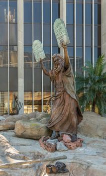 Garden Grove, California, USA - December 13, 2018: Crystal Christ Cathedral. Bronze statue of Moses putting the ten commandments of two tables in the air. Some green foliage, Tower of hope in back. Cathedral reflected.