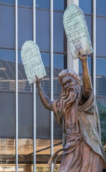 Garden Grove, California, USA - December 13, 2018: Crystal Christ Cathedral. Closeup of Bronze statue of Moses putting the ten commandments of two tables in the air. Some green foliage, Tower of hope in back.