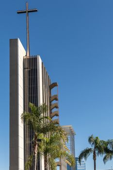 Garden Grove, California, USA - December 13, 2018: Crystal Christ Cathedral. Tower of Hope with large cross against blue sky. Green palm trees and golden bell display in back.