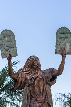 Garden Grove, California, USA - December 13, 2018: Crystal Christ Cathedral. Closeup of Bronze statue of Moses putting the ten commandments of two tables in the air. Some green foliage, light blue sky.