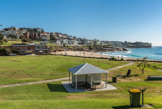 Sydney, Australia - February 11, 2019: Green Bronte Park with sandy beach and blue Tasman Sea. Buildings on North Shore and Bronte Surf Life Saving Club house.